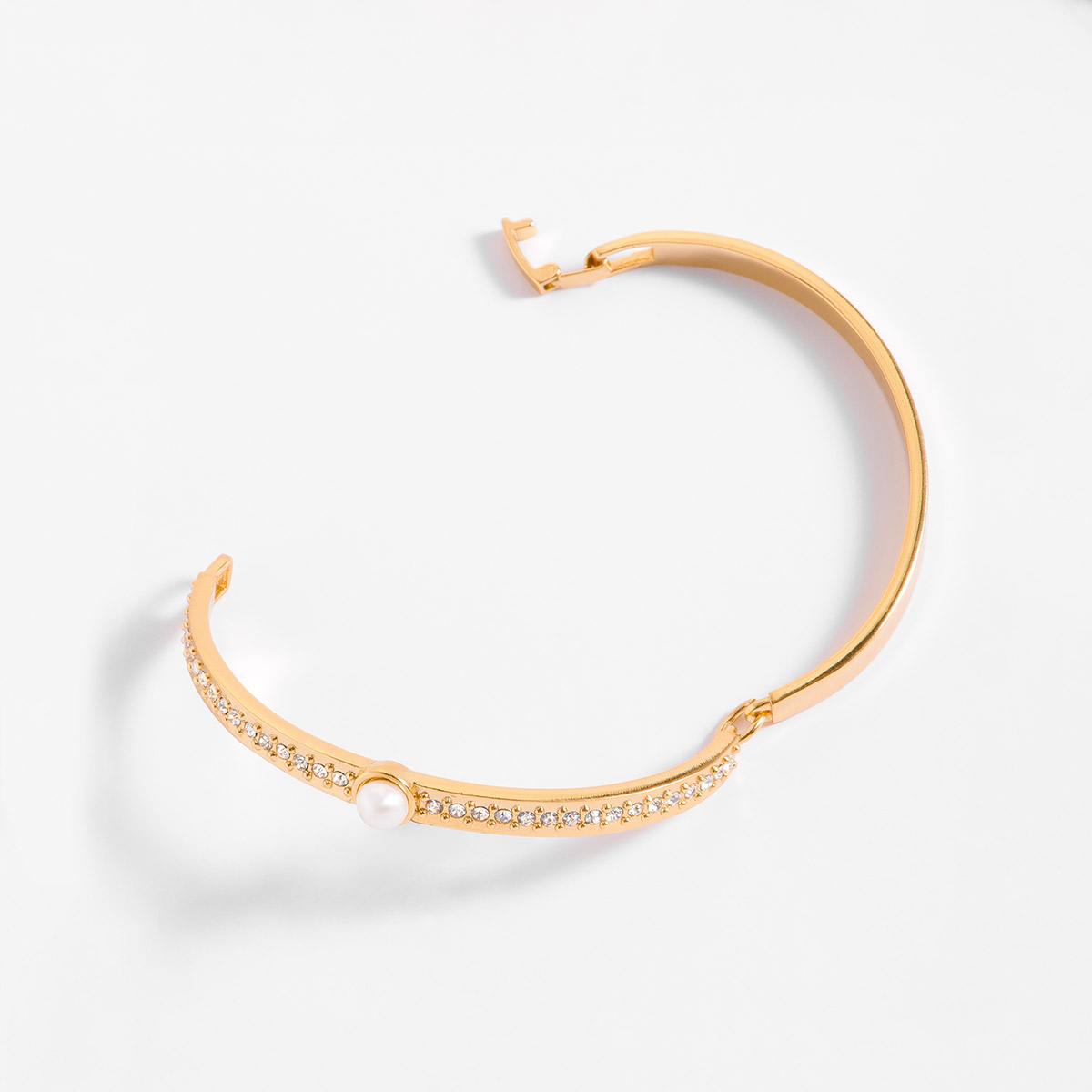 Sofisticado brazalete ajustable en baño de oro 18k, con un diseño especial; al centro una perla en tono crema, acompañada a los lados con piedras en tono cristal. Combínalo con tu collar y aretes.
-        Pulsera
-        Medida Ajustable
-        Baño de Oro 18k
-        Perla en tono crema
-        Piedras en tono cristal
